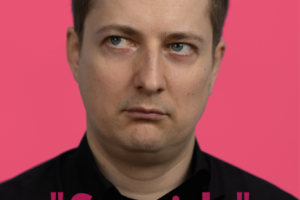 Vīrietis melnā kreklā ar nedaudz nopietnu sejas izteiksmi skatās sāniski pa kreisi uz rozā fona. Attēlā redzams teksts: "Leimaņu Tautas namā", ""Smaids"", "Riktīgi pozitīva stand-up komēdija", "Sestdien/28. martā/plkst.15.00", "Komikis: Maksims Trivaškevičs", "ieeja: 3,00 EUR".