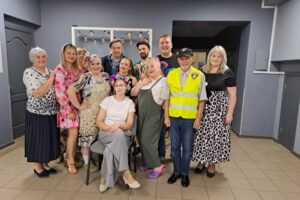 Attēls ar Biržu Tautas nama Amatierteātri &quot;BIRZĪTE&quot;, ar aptuveni 16 cilvēkiem, vīriešiem un sievietēm dažādos vecumos, pozē telpās, smaidot kamerā. Daži no viņiem ir tērpušies kostīmos, kas liecina par teātra izrādi vai priekšnesumu, piemēram, sieviete ar ziediem matos un priekšautu, un vīrietis dzeltenā atstarojošā vestē.