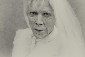 Liepājas Teātris. Melnbalta fotogrāfija ar aktieri smagā teātra grimmā, kurā attēlots gados vecs cilvēks ar baltiem matiem un baltu plīvuru, kas skatās tieši uz skatītāju. Liepājas teātra viesizrāde Krustpils kultūras centrā 22. aprīlī plkst. 19.00. Mārtiņš Makdona. Līnenas skaistumkaraliene, skumja komēdija. Lomās: Inese Kučinska, Mārtiņš Kalita, Edgars Ozoliņš, Artūrs Irbe. Režisors – Dž.Dž.Dzilindžers.